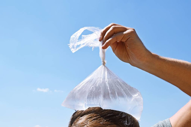 Un uomo sulla ventina si raffredda la testa con un sacchetto di plastica pieno di acqua fredda di fronte a un cielo blu chiaro senza nuvole - Raffreddamento rinfrescante nella calda estate in Europa
  - Foto, immagini