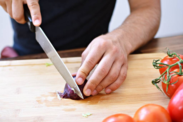 A person cuts a red onion on a wooden board for a fresh crunchy healthy salad - close-up on the hands of the person - concept for healthy nutrition with fresh organic vegetables - Fotó, kép
