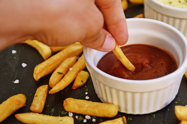  fresh potato fries of golden yellow fried in a bowl full of ketchup, against a dark marble background, on which fries and salt grains and spices lie - Φωτογραφία, εικόνα