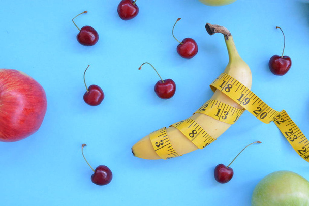 Diferentes tipos de frutas, como manzanas, cerezas y plátanos se encuentran sobre una base de un solo pie y se envuelven en una cinta métrica - concepto para la pérdida de peso saludable que se burla
 - Foto, imagen