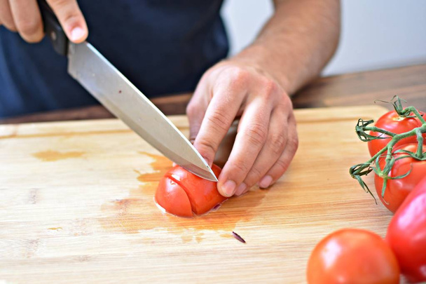 A person cuts a tomato on a wooden board for a fresh crunchy healthy salad - close-up on the hands of the person - concept for healthy nutrition with fresh organic vegetables  - Zdjęcie, obraz