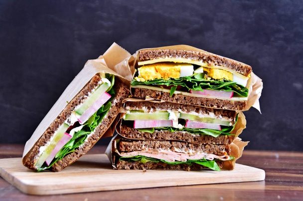 Various sandwiches of sunflower seed bread with cheese, sausage, egg and salad as a healthy diet are stacked on a dark background  - Photo, Image