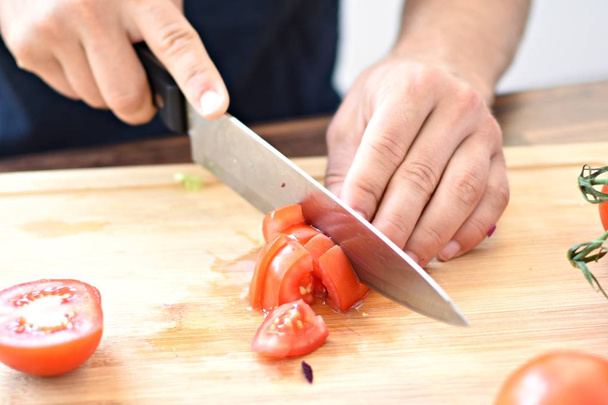A person cuts a tomato on a wooden board for a fresh crunchy healthy salad - close-up on the hands of the person - concept for healthy nutrition with fresh organic vegetables  - Valokuva, kuva