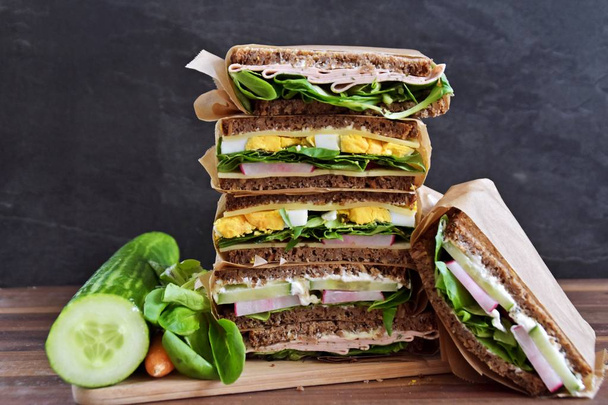 Várias sanduíches de pão de semente de girassol com queijo, salsicha, ovo e salada como uma dieta saudável são empilhadas em um fundo escuro, ao lado de uma fita métrica como uma alusão à aptidão
  - Foto, Imagem