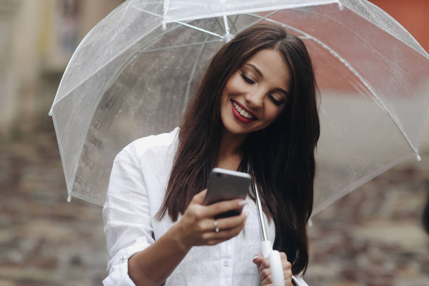 Close up portrait of smiling beautiful girl with umbrella, using smartphone, red wall on the background. - Фото, изображение