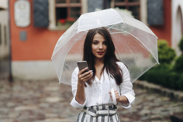Portret marzycielski piękna dziewczyna z parasolem, za pomocą smartfona, czerwony dom na tle. Mała głębia ostrości. - Zdjęcie, obraz