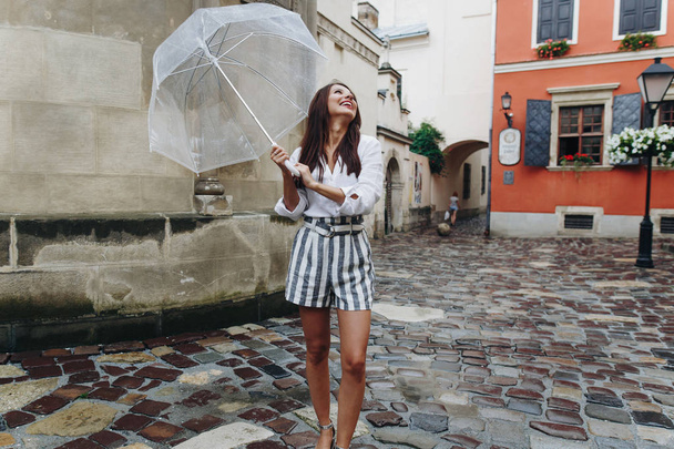 Spaziergang in der Innenstadt, junge, modische Frau, die durch die Stadt läuft, Gesichtsausdruck. schöne brünette junge Frau in kurzen Hosen, die mit durchsichtigem Regenschirm auf der Straße spaziert. - Foto, Bild