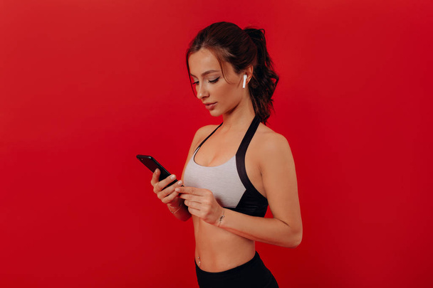 Foto de cerca de una chica atlética con cabello oscuro que usa traje deportivo escuchando música y usando un teléfono inteligente sobre un fondo aislado
  - Foto, imagen