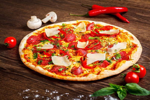 Pizza Italian with parmezan cheese, tomatoes, salami, pepperoni and green basil on a brown table decorated by mushrooms, red sweet pepper and cherry tomatoes - Photo, Image
