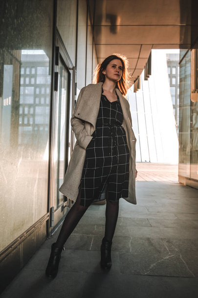 Photo of a girl in a coat and dress against the background of a modern business center, backlight - Фото, зображення
