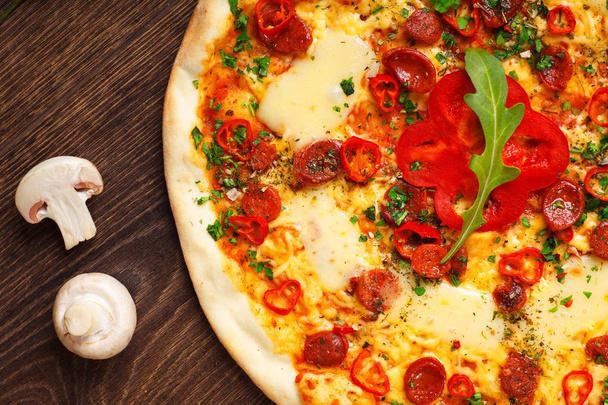 Close-up Italiaanse pizza met gesmolten kaas, worst, snack sticks, peper en verse groene oregano bladeren op een bruine tafel versierd met champignons, rode paprika en cherry tomaten - Foto, afbeelding