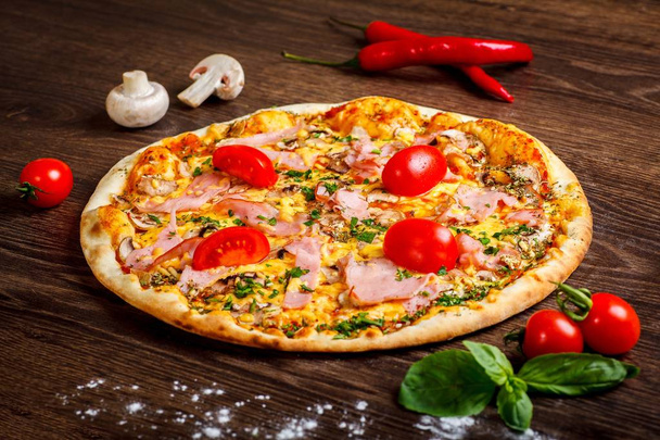 Pizza Italian with melted mozzarella cheese, red cherry tomatoes, ham and fresh green herbs on a brown table decorated by mushrooms, red sweet pepper and cherry tomatoes - Photo, Image