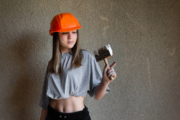 Une fille tient un pinceau de construction pour peindre un mur en béton. Elle porte un casque orange protecteur
. - Photo, image