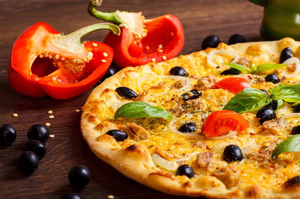 Close-up vegetarische tonijn Italiaanse pizza gesmolten kaas, zwarte olijven, rode cherry tomaten en verse groene Basilicum bladeren op een bruine tafel versierd met champignons, zwarte olijven, rode peper en verse rucola - Foto, afbeelding