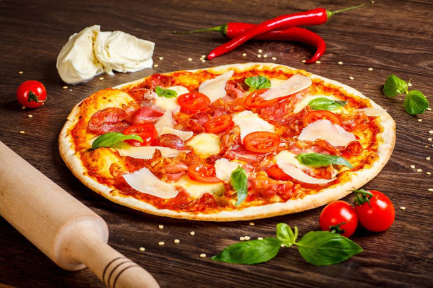 Pizza italiana con mozzarella fusa, parmezan, salame, pomodorini rossi e foglie di basilico verde fresco su un tavolo marrone decorato con mozzarella, peperone rosso dolce, ciliegia e mattarello
 - Foto, immagini