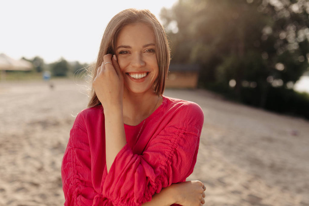 hübsche Frau mit sanftem Lächeln, die vor dem Hintergrund des Sandstrandes im Sonnenlicht in die Kamera blickt  - Foto, Bild