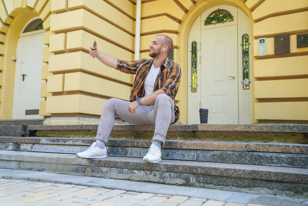 joven buen aspecto chico barbudo audaz está sentado al aire libre en las escaleras en frente de su casa haciendo selfie y sonriendo. tiene auriculares en los oídos. vistiendo camisa amarilla y jeans
. - Foto, Imagen