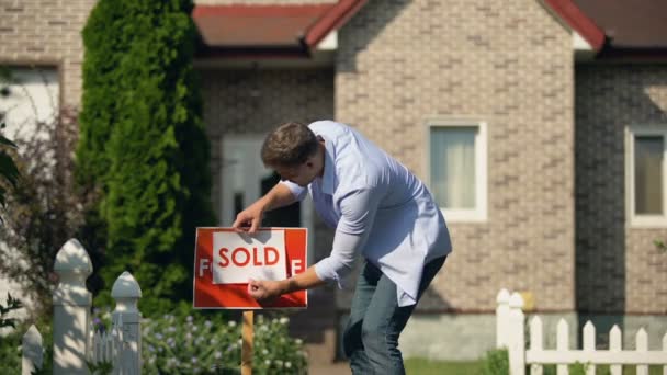 Proud man changing for sale to sold signboard in front of house, buy new home - Video