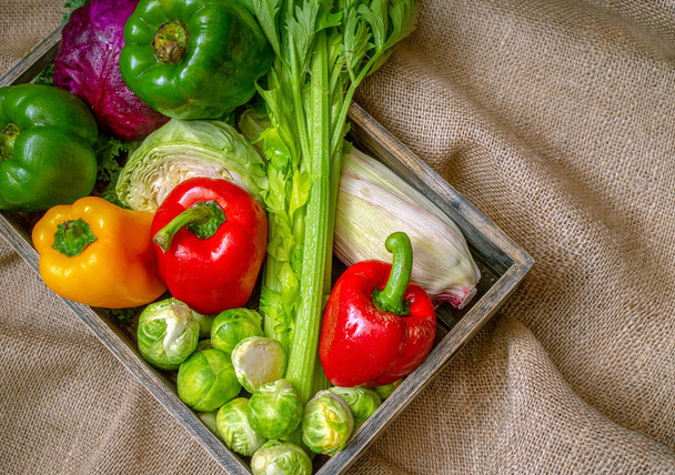 Különböző nem mérgező friss zöldségek kerülnek egy fadobozban helyezni a szőnyegen. Sok zöldség lila és zöld hajtások, piros paprika, sárga és zöld, kukorica és mások - Fotó, kép