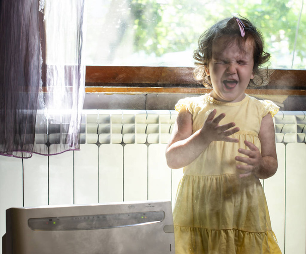 Ένα κοριτσάκι σε ένα σκονισμένο δωμάτιο. Καθαριστής αέρα και βήχας παιδί. Σκόνη - Φωτογραφία, εικόνα