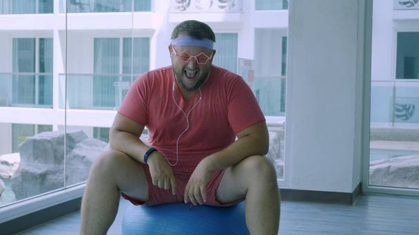 Playful fat man in a pink T-shirt and pink glasses is engaged in fitness with dumbbells and a fit ball in the gym. man listening to music on headphones. - Φωτογραφία, εικόνα