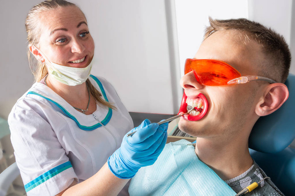 Tandheelkunde. Vrolijke vrouwelijke tandarts en jonge mannelijke patiënt. Onderzoek van de tanden met behulp van een tandheelkundige spiegel close-up. - Foto, afbeelding