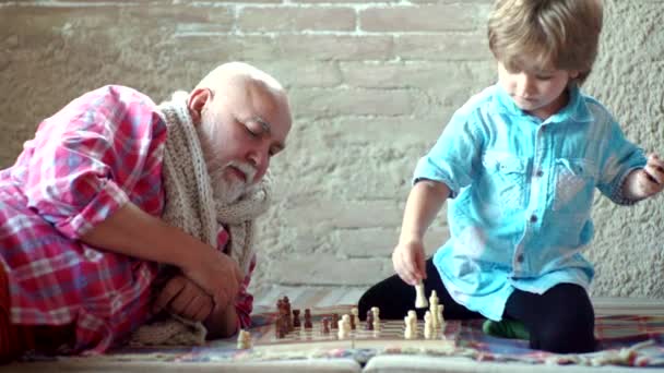 Yakışıklı büyükbaba ve torun evde birlikte vakit geçirirken satranç oynuyorlar. Küçük çocuk büyükbabasıyla satranç oynuyor. Küçük çocuk satranç oynayan olgun adam. - Video, Çekim