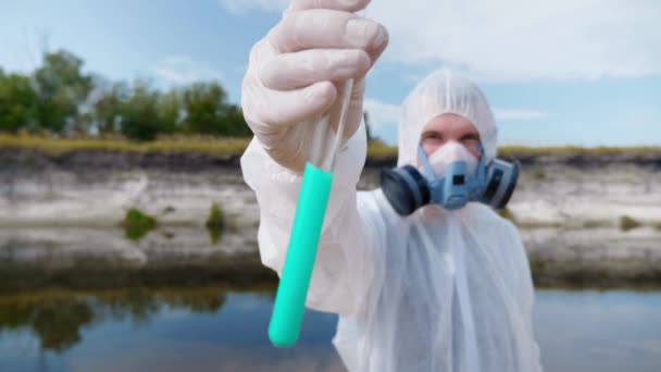 A man in a protective suit and a respirator shows a test tube with analysis of water from polluted river or lake. Scientist takes samples in vitro. Ecological disaster. Save the planet. 4K footage. - Footage, Video