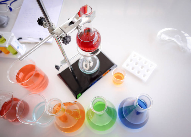 ガラス検査用の赤い液体がラックにフラスコを積み込みます。オレンジ、青、緑、赤の化学実験室のガラス製品の下のアルコルホールランプの場所 - 写真・画像
