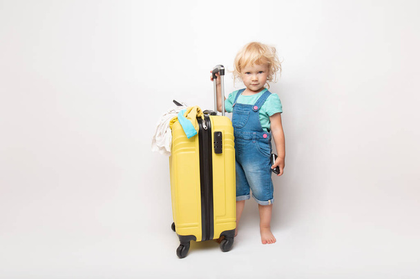 Funny curly little baby girl in a hat, T-shirt and jeans with a yellow suitcase is smiling on white background waiting for an airplane. concept trip with kids. - Photo, image