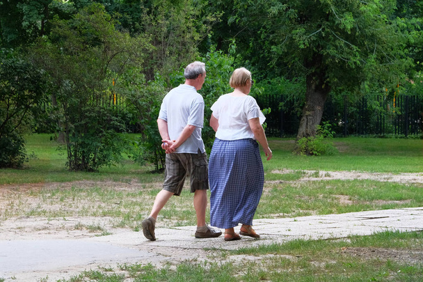 Elderly couple is walking in green city park on day off. Middle aged couple is walking. Green trees on sides of trail. - Photo, Image