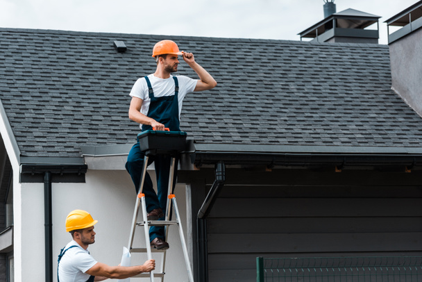 handsome repairman sitting on roof and holding toolbox near coworker in helmet  - Photo, Image