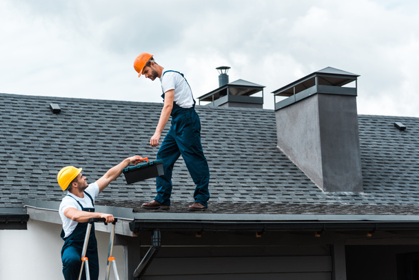 handyman in helmet giving toolbox to handsome colleague standing on rooftop  - Photo, Image