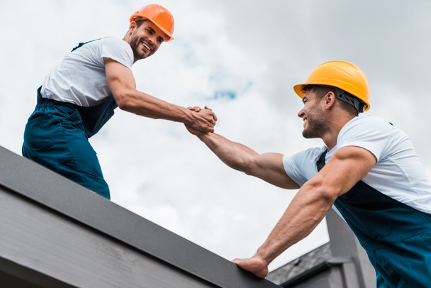 low angle view of cheerful handymen in helmets holding hands  - Photo, Image