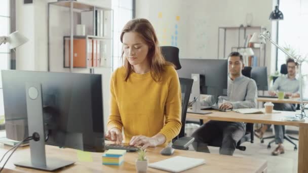 Beautiful and Smart Red Haired Female Specialist Sitting at Her Desk Works on a Desktop Computer. In the Background Modern Bright Office with Diverse Group of Professionals Working for Growing Startup - Metraje, vídeo