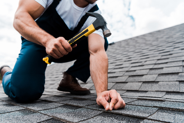 cropped view of handyman in uniform holding hammer while repairing roof  - Φωτογραφία, εικόνα