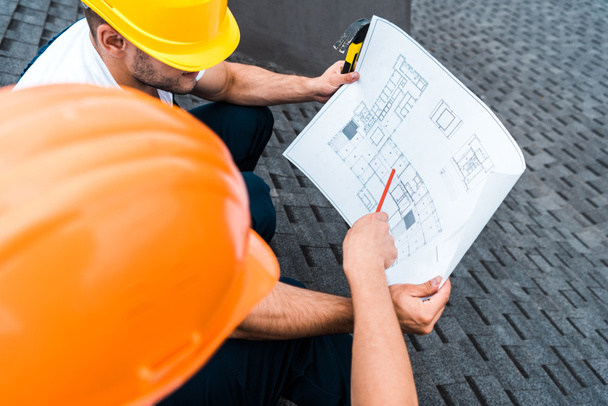 selective focus of architect in helmet holding pencil near blueprint and coworker  - Photo, Image