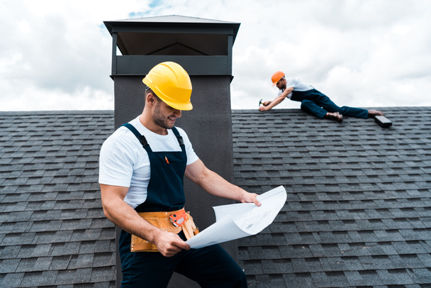 селективный фокус красивого строителя в шлеме, держащего чертеж рядом с коллегой, ремонтирующим крышу
  - Фото, изображение