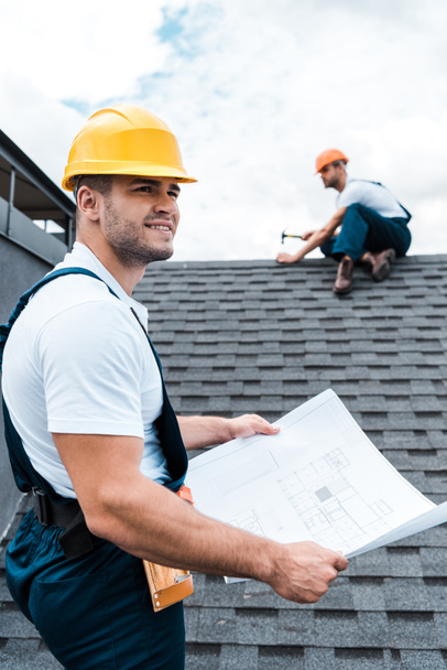 Selektywny fokus przystojny budowniczy w kask gospodarstwa planu w pobliżu kolega naprawy dachu  - Zdjęcie, obraz