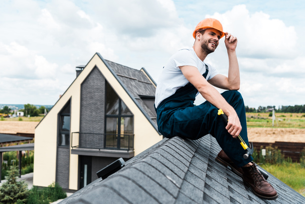 gelukkige klusjesman in oranje helm zittend op dak en hamer houden  - Foto, afbeelding