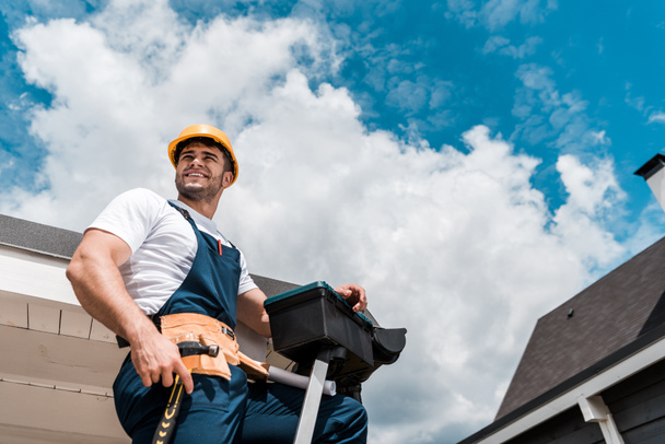 low angle view of happy repairman in helmet holding hammer and toolbox - Photo, Image