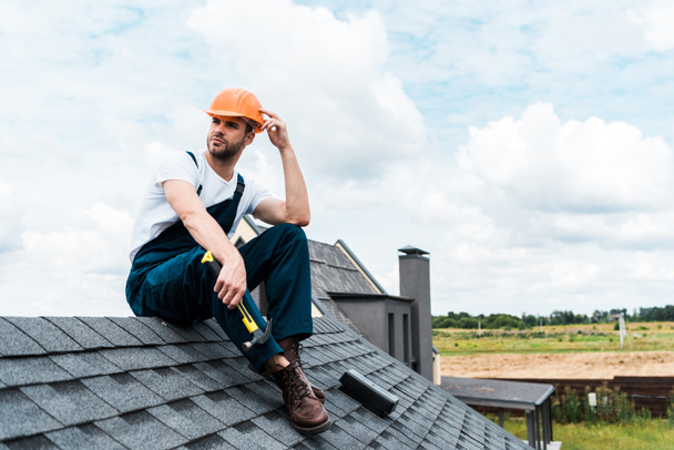 handsome handyman in orange helmet sitting on roof and holding hammer  - Photo, Image