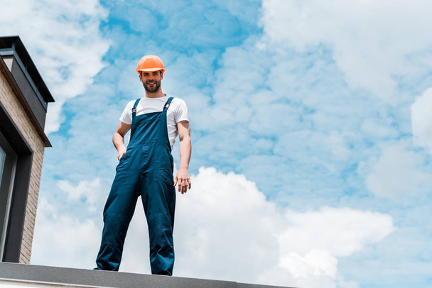 low angle view of happy repairman in helmet and uniform standing against blue sky with clouds  - 写真・画像