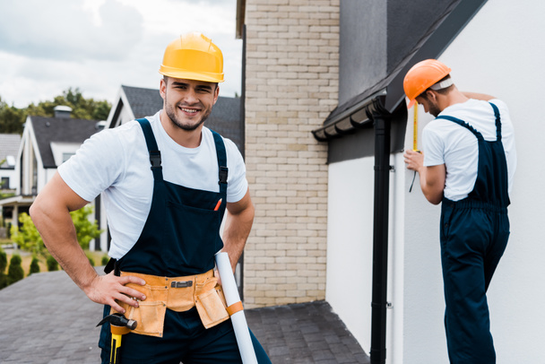 selective focus of cheerful repairman standing with hands on hips and smiling near coworker  - Photo, Image