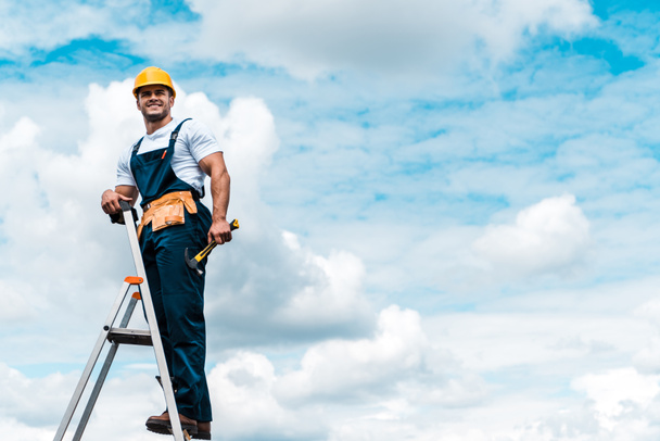 riparatore allegro in piedi sulla scala e sorridente contro il cielo blu con le nuvole
  - Foto, immagini
