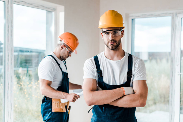 selective focus of handsome handyman in goggles standing with crossed arms near coworker  - Photo, Image