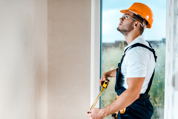 handsome repairman in goggles holding measuring tape and looking at wall near window  - Foto, Bild