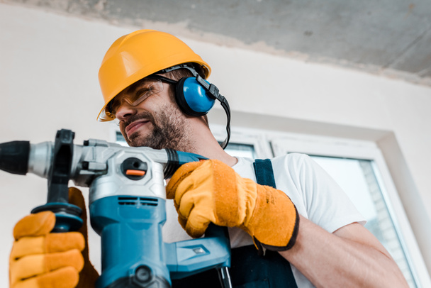 selective focus of handyman in helmet and yellow gloves using hammer drill  - Foto, Imagem