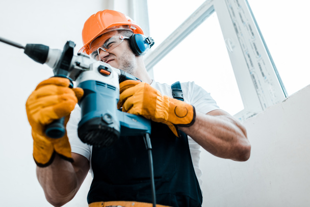 low angle view of handyman in helmet and yellow gloves using hammer drill  - 写真・画像