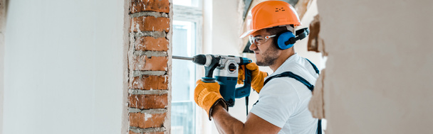 panoramic shot of handyman in uniform and yellow gloves using hammer drill  - Foto, afbeelding